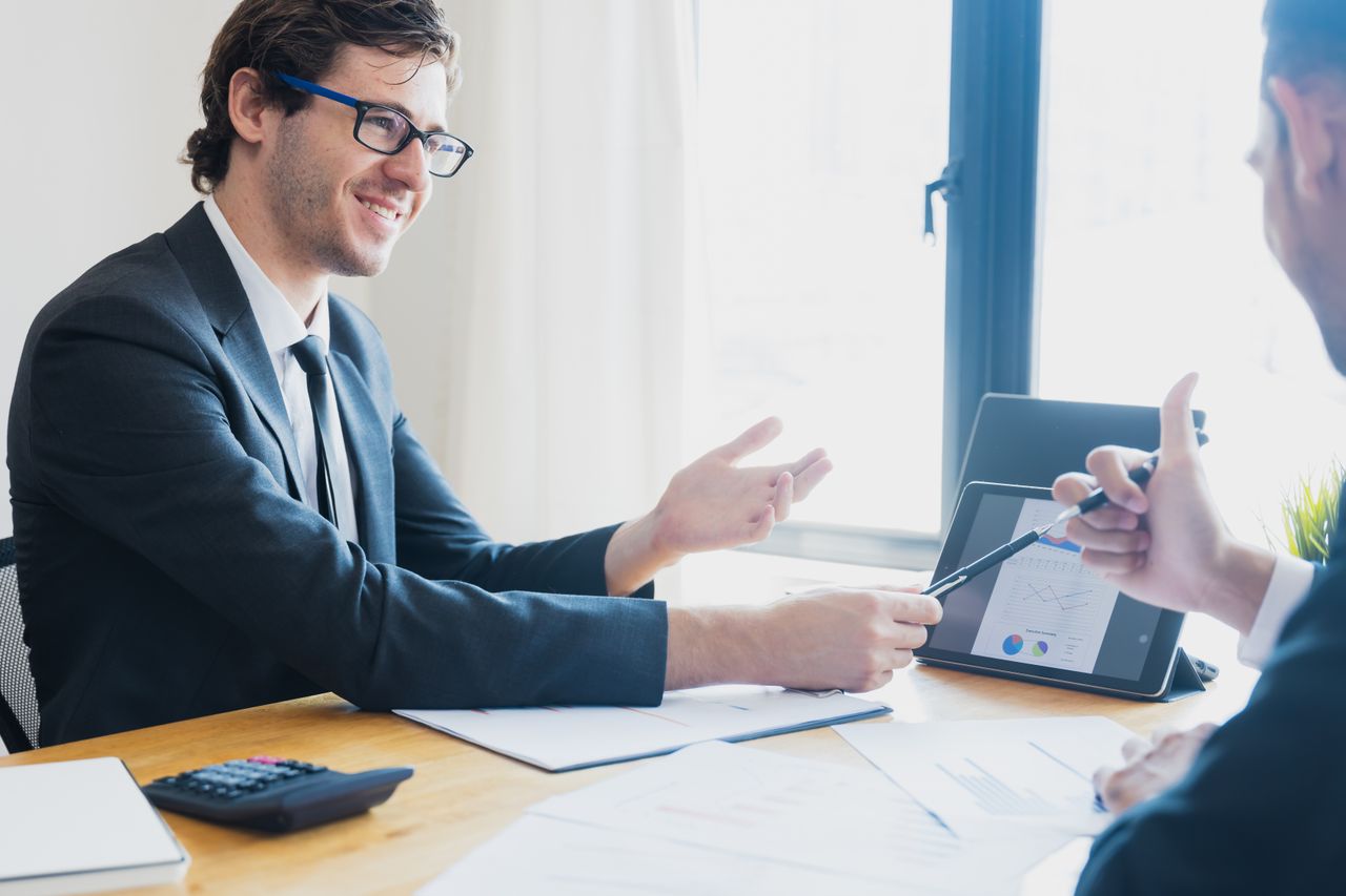 team-03 Two businessmen in formal attire discussing a business report in an office setting with a tablet.